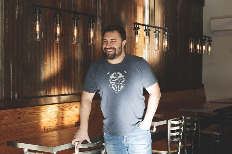 A smiling man in a graphic tee standing in the wood-paneled Iron Tap Station taproom