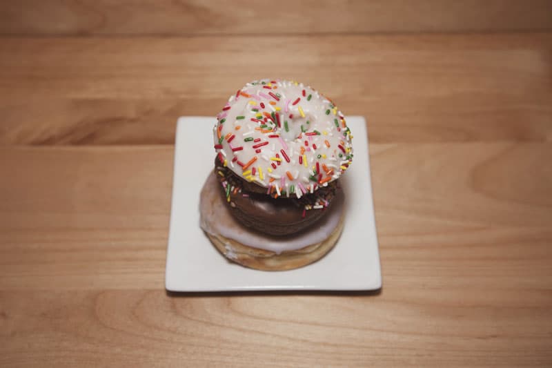 Two Coco Donuts donuts, one with rainbow sprinkles and one chocolate-glazed, on a plate on a wood table