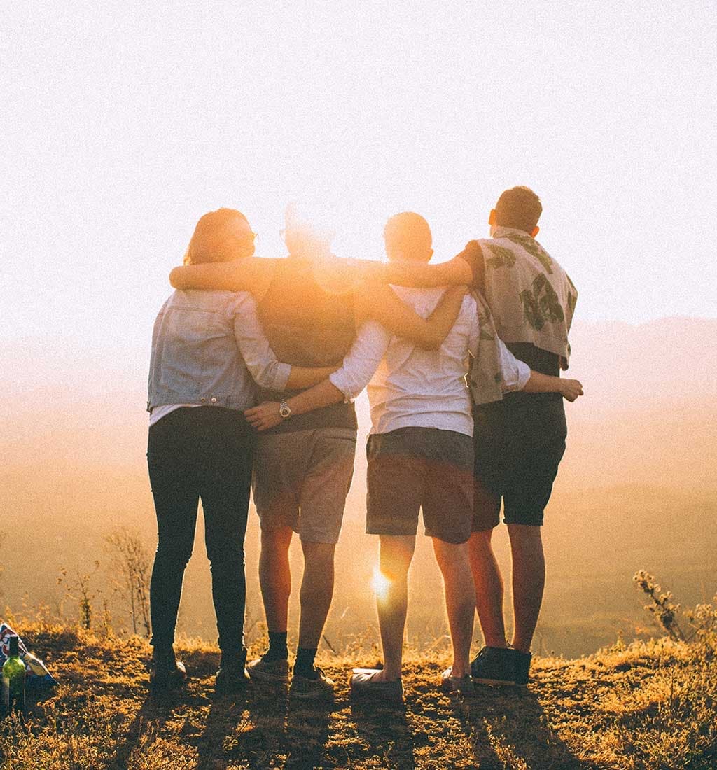 Four friends with arms around each other watching the sun set over a hillside