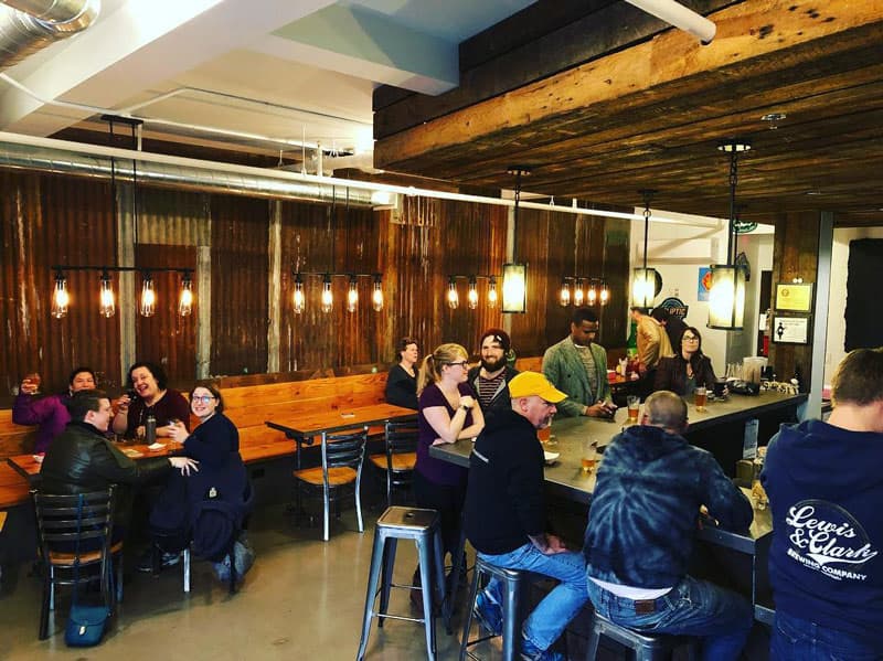 Iron Tap Station taproom filled with people at the bar and tables under wood beams and Edison bulbs