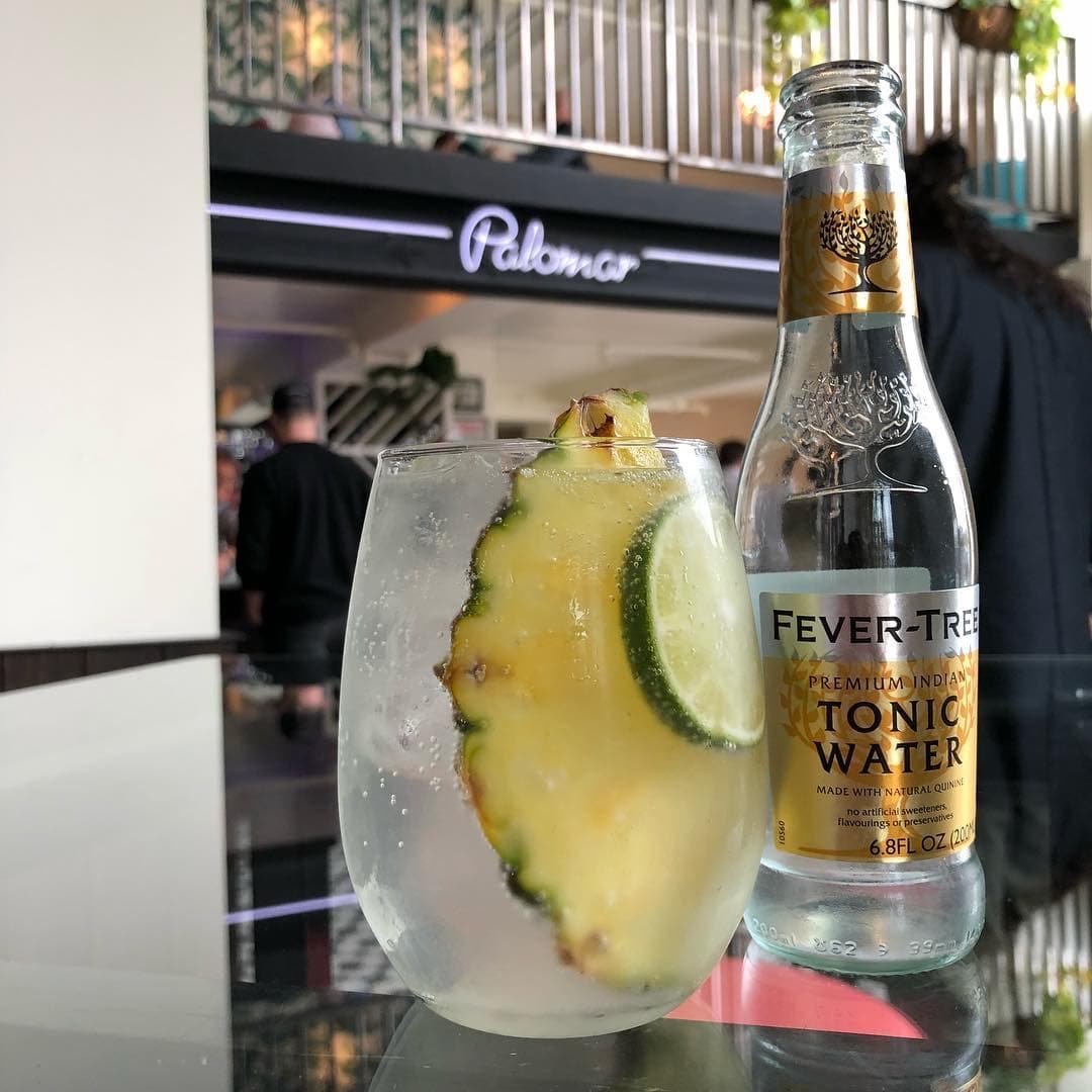 Palomar cocktail with a pineapple wedge and lime beside a tonic water bottle, the bar's sign behind