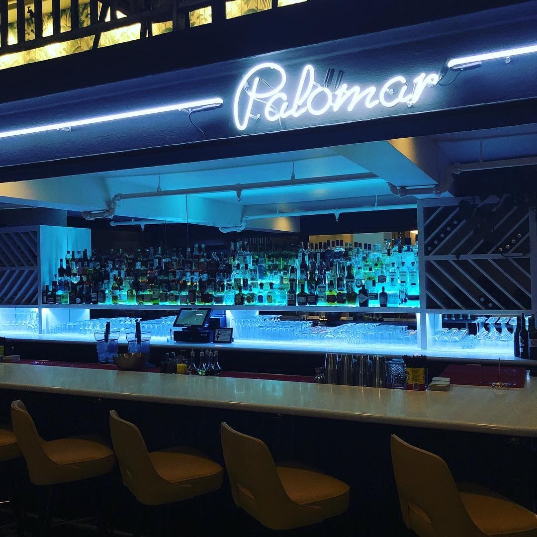 Palomar's neon sign glowing above a blue-lit, well-stocked bar and yellow bar stools